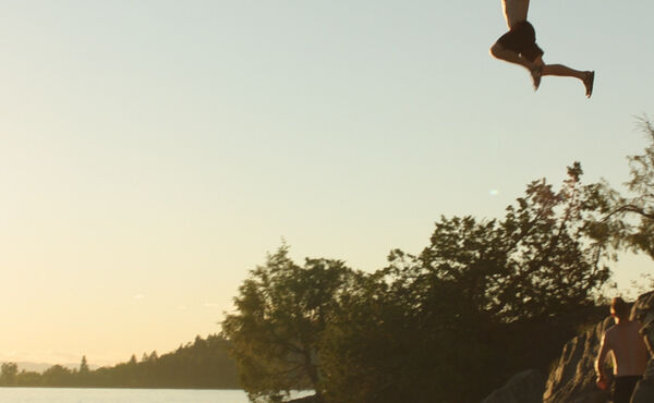 Person jumping off rocks into a lake at sunset, with trees and friends nearby.